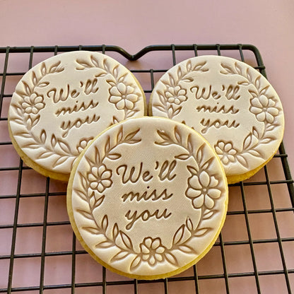 Three cookies with 'We'll miss you' text on a cooling rack against a pink background