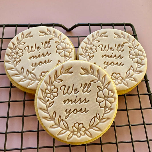 Three cookies with 'We'll miss you' text on a cooling rack against a pink background