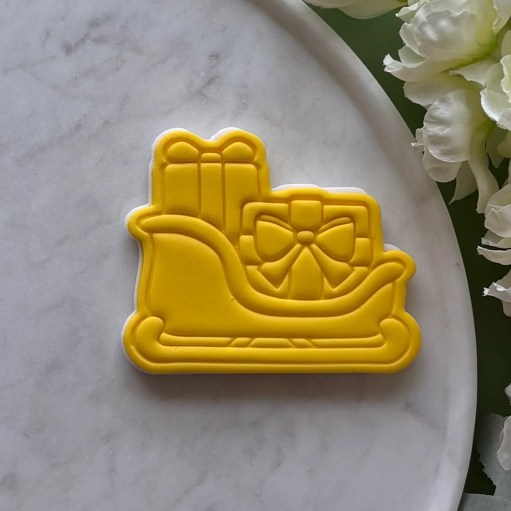 Yellow sled cookie cutter with a bow on a marble surface with flowers.