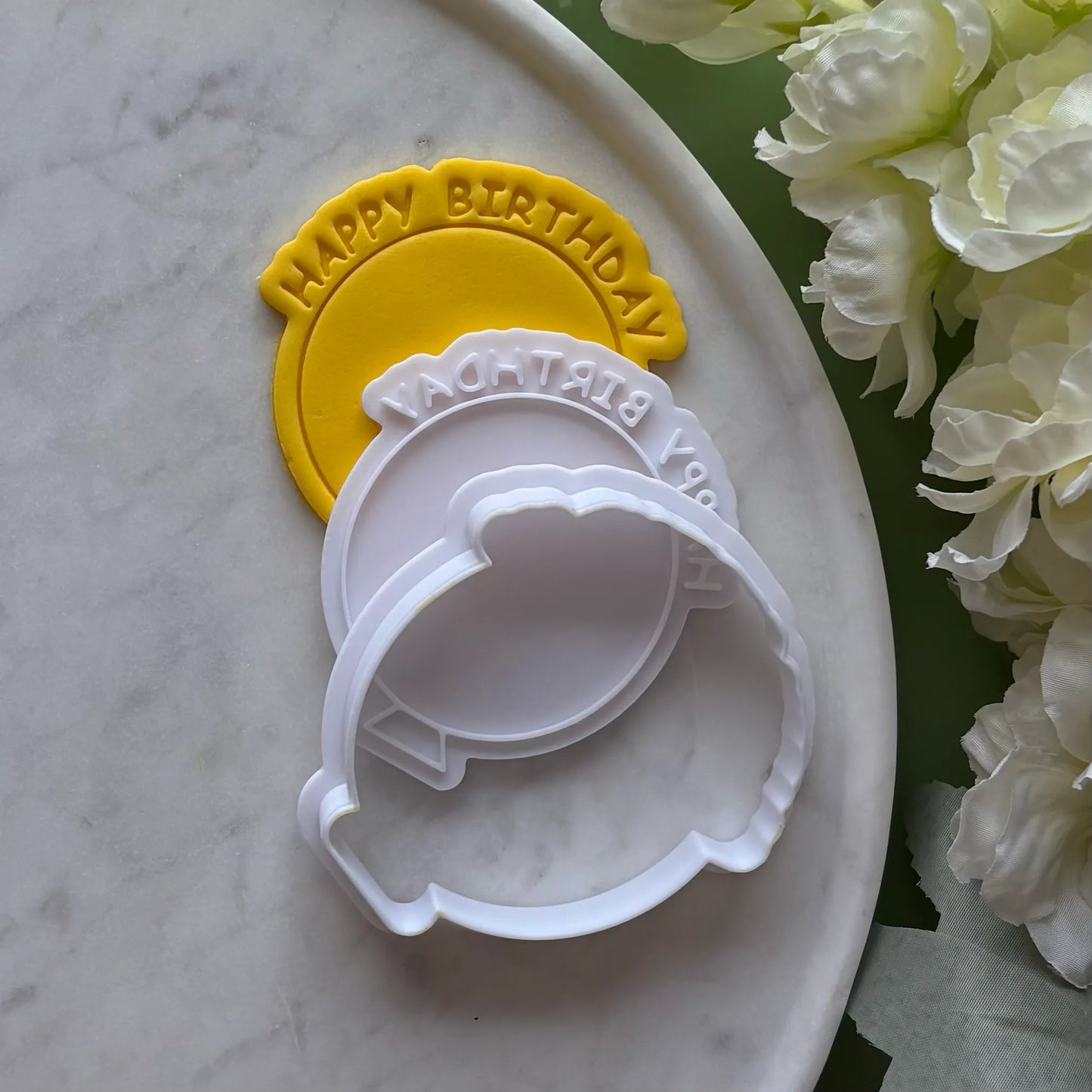 Two cookie cutters with 'Happy Birthday' text on a marble surface with flowers.