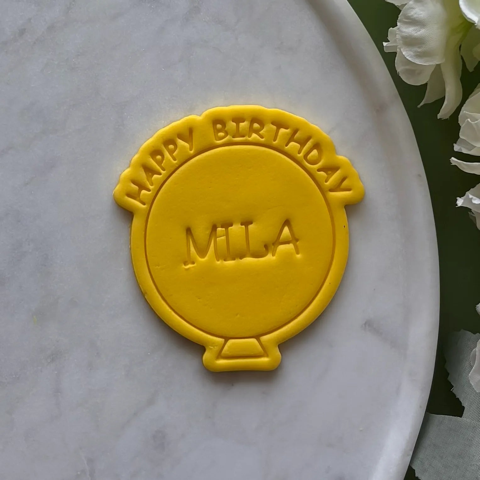 Yellow 'Happy Birthday Mila' cookie on a marble surface with flowers.