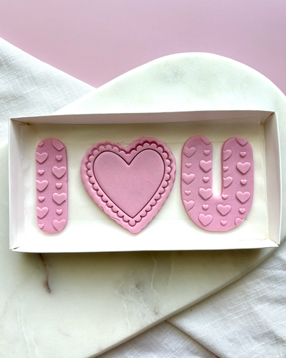 Pink cookie cutters shaped like 'I ❤️ U' on a white marble surface with a pink background.