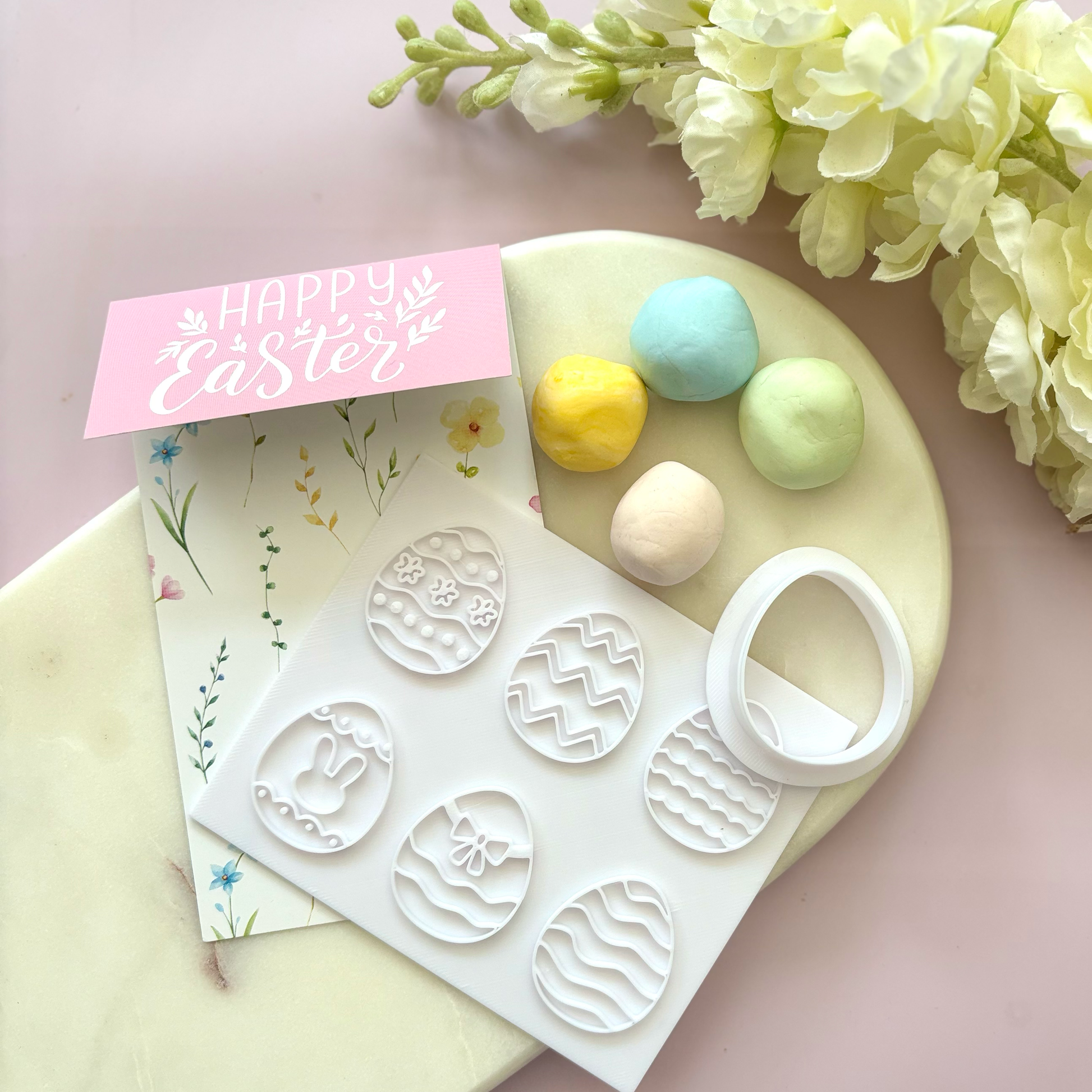Easter-themed cookie cutters with pastel-colored eggs and a 'Happy Easter' card on a pink surface.