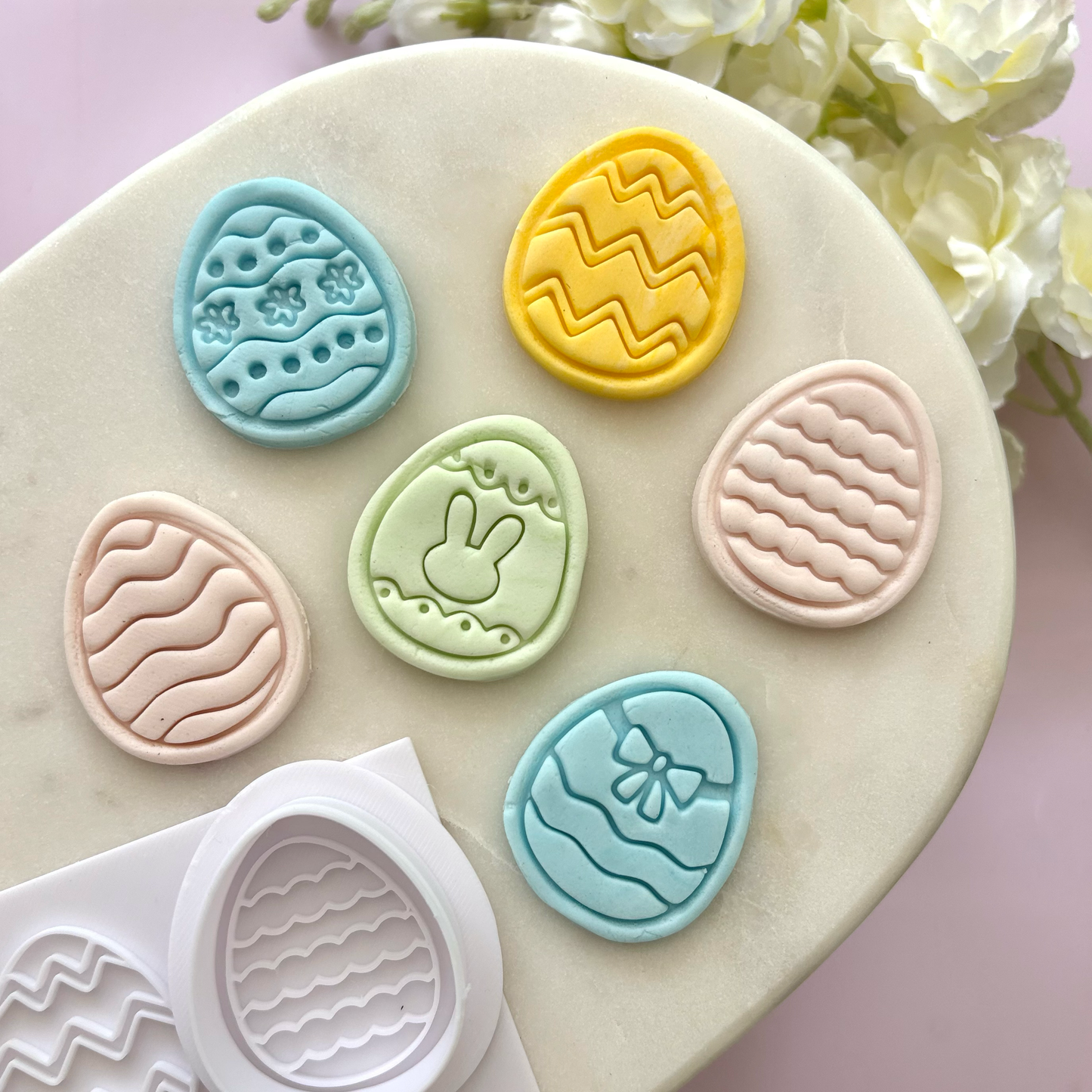 Set of colorful Easter egg-shaped cookie cutters on a marble surface with flowers in the background.