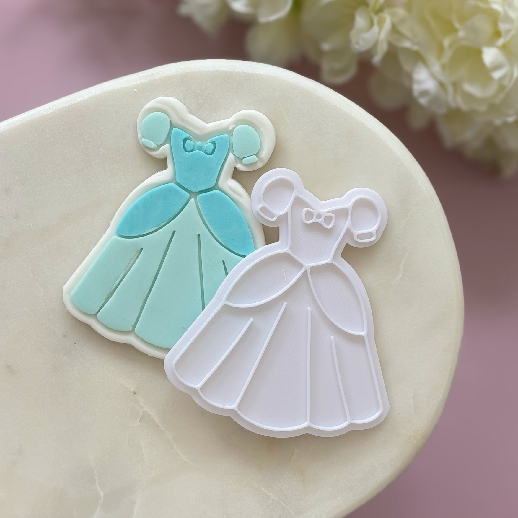 Two dress-shaped cookie cutters, one blue and one white, on a marble surface with flowers in the background.