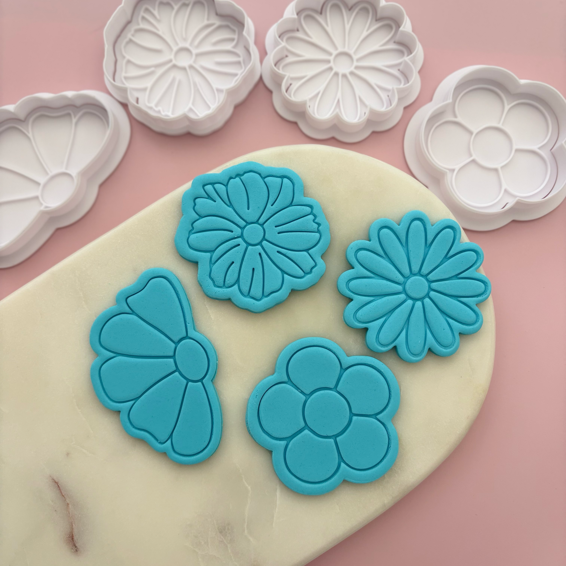 Blue flower-shaped cookie cutters on a pink surface with white flower-shaped cookie cutters in the background.