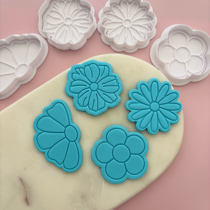 Blue flower-shaped cookie cutters on a pink surface with white flower-shaped cookie cutters in the background.