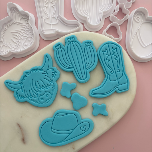 Set of blue cookie cutters shaped like a highland cow, cactus, cowboy boot, and hat on a pink background.
