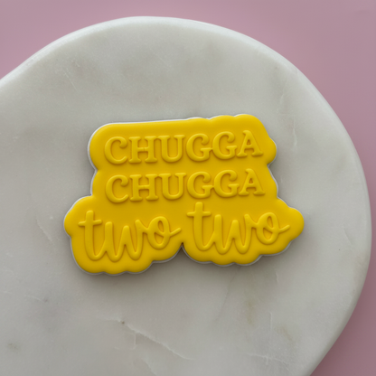 Yellow cookie cutter with 'CHUGGA CHUGGA two two' text on a white marble surface.