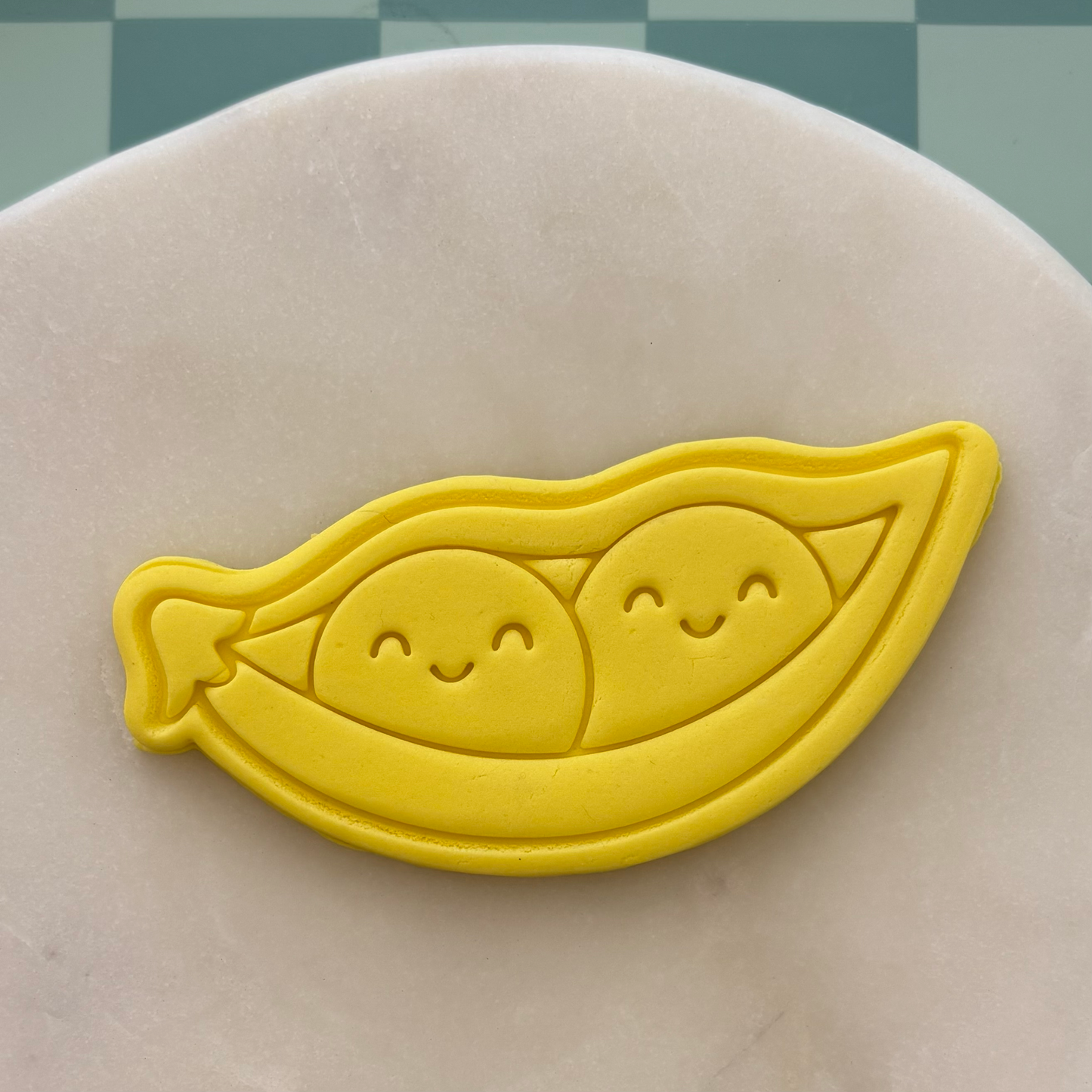 Yellow cookie cutter shaped like a pod with two smiling peas on a gray surface.