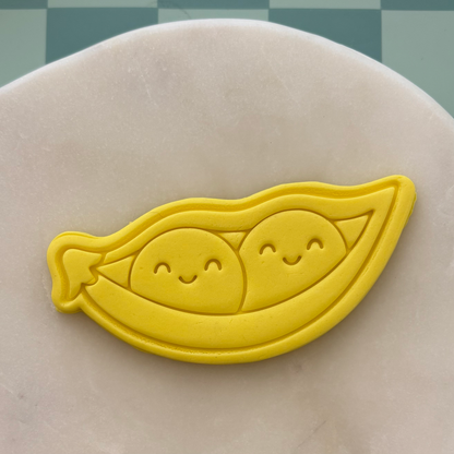 Yellow cookie cutter shaped like a pod with two smiling peas on a gray surface.