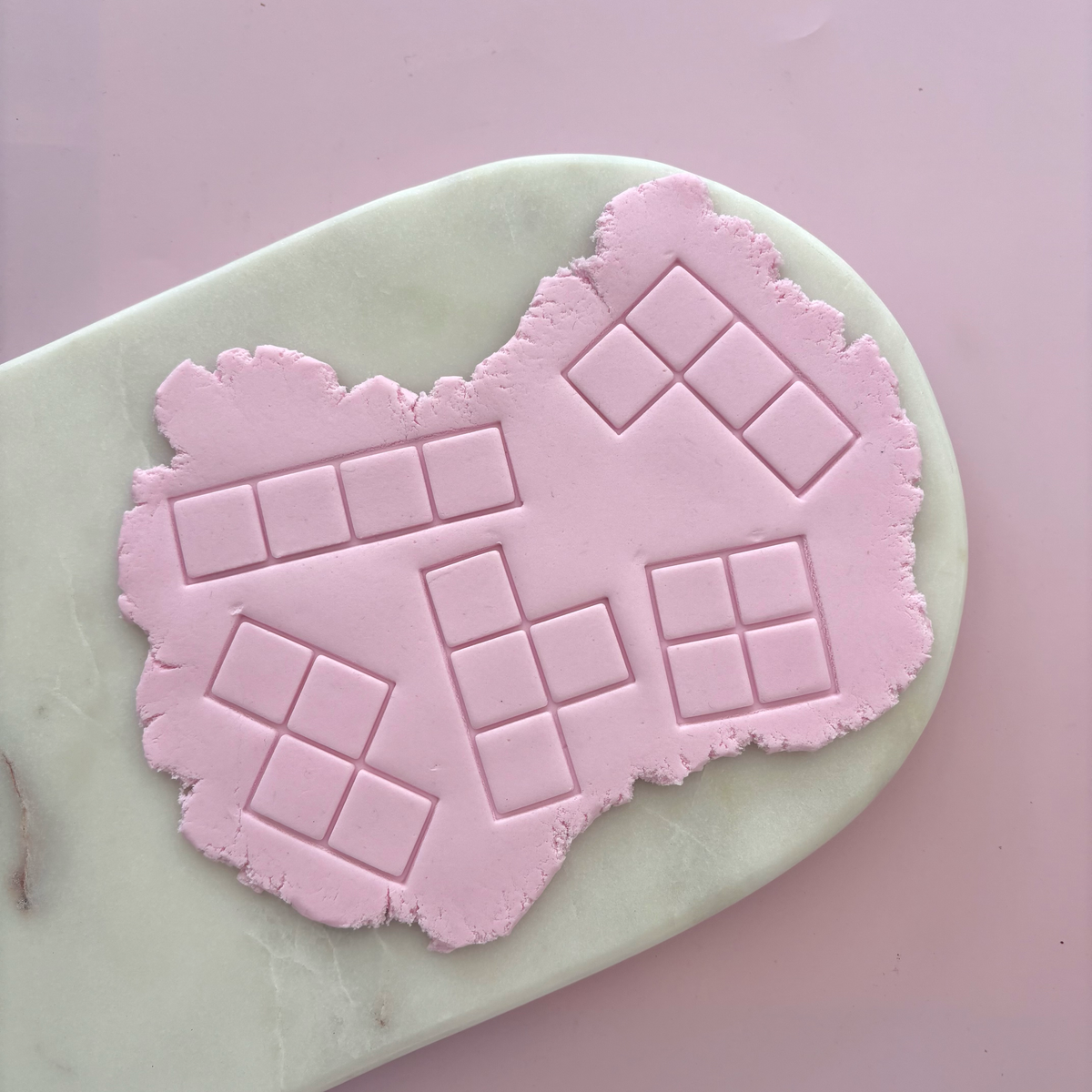 A pink silicone baking mat with embossed square grid patterns, shown on a pale green marble board against a solid pink background.