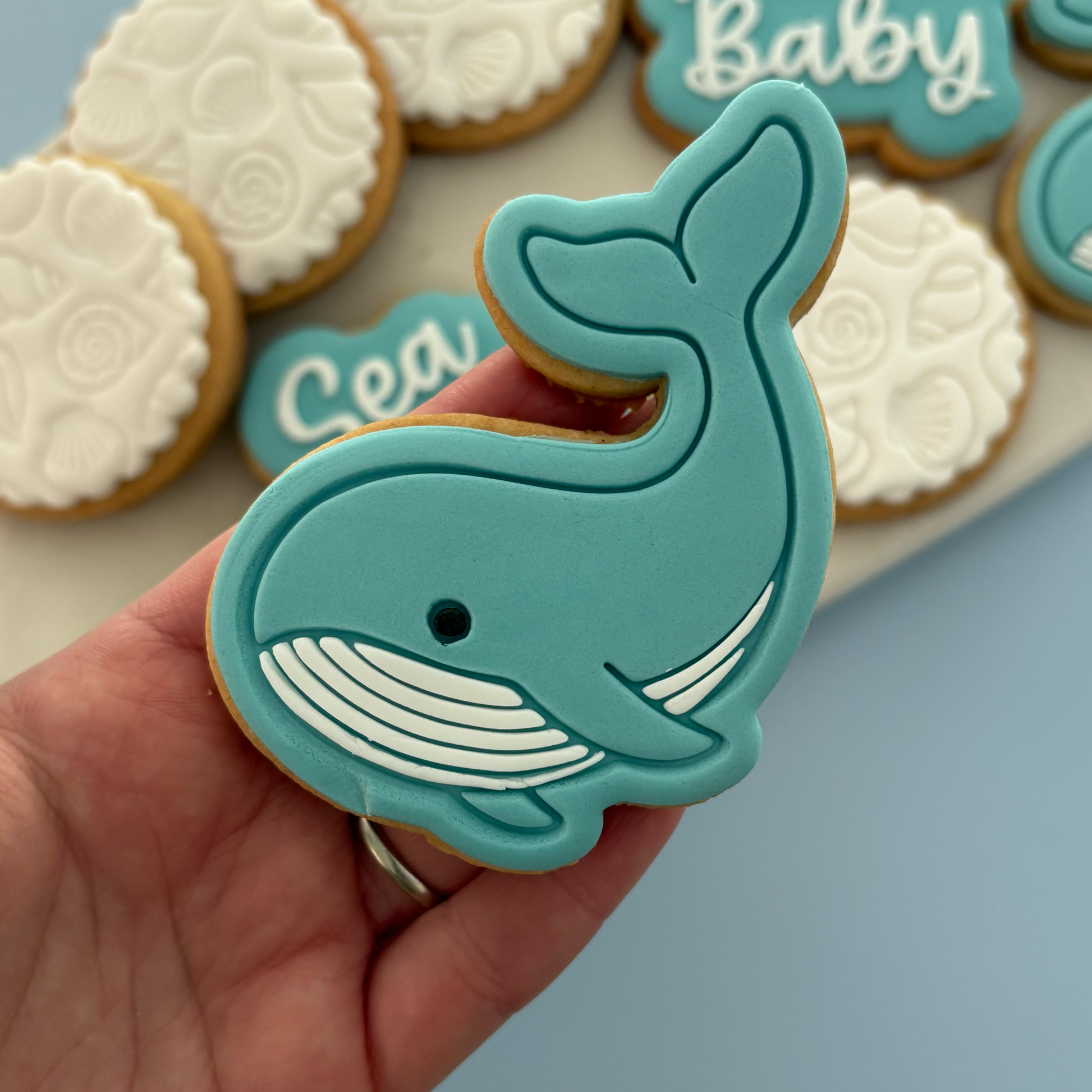 A hand holds a whale-shaped decorated cookie in front of other iced cookies on a tray against a soft blue background.