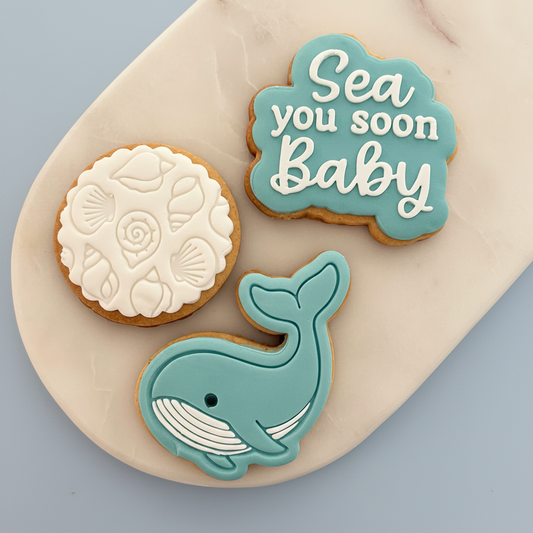 Three decorated round cookies arranged on a light beige marble board against a soft blue-gray background. One cookie is turquoise with white script reading 'Sea you soon Baby,' one is white with shell motifs, and one is turquoise with a whale design.