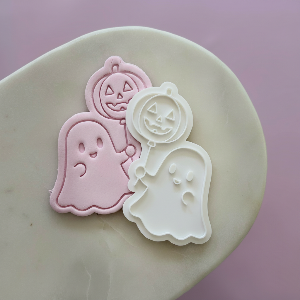 Halloween-themed cookie cutters or stamps on a beige stone surface against a soft pink background: a pink ghost, a white ghost, and a pink pumpkin with a jack-o'-lantern face.