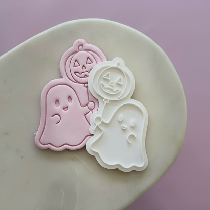 Halloween-themed cookie cutters or stamps on a beige stone surface against a soft pink background: a pink ghost, a white ghost, and a pink pumpkin with a jack-o'-lantern face.