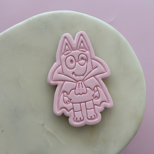 A pink fondant cutter stamp shaped like a cartoon  shown pressed into a pale cream-colored clay disc against a soft pink background.