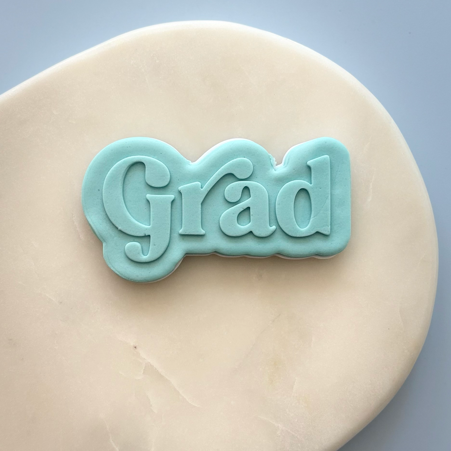 A light blue fondant cookie cutter or stamp shaped like the word 'Grad' in cursive, displayed on a beige fondant backdrop against a soft blue background.