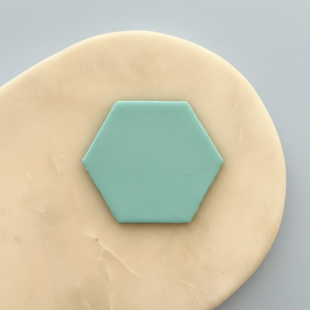 A small mint green hexagonal tile or coaster centered on a beige clay or dough disc against a light gray background.