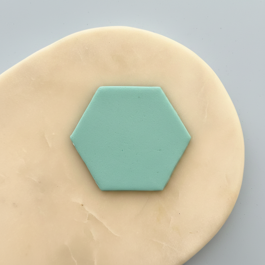 A small mint green hexagonal tile or coaster centered on a beige clay or dough disc against a light gray background.