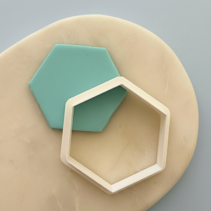 A hexagonal cookie cutter in a warm cream color resting on a beige marble surface next to a mint green hexagonal clay cutout, set against a solid gray-blue background.