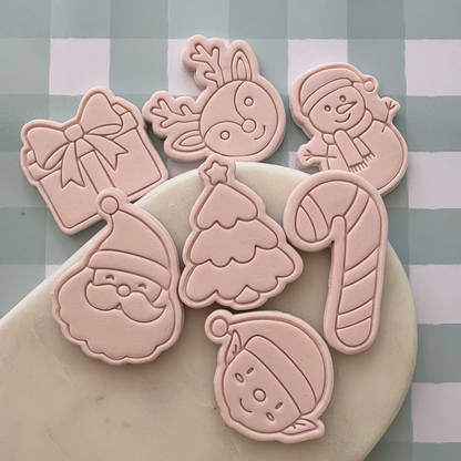 A set of six pale pink Christmas-themed cookie cutters or stamps arranged on a white marble board over a green and white checkered surface. The shapes include Santa Claus, a Christmas tree, a candy cane, a snowman, a reindeer, a gift box, and a ornament.