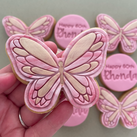 A hand holds a butterfly-shaped sugar cookie decorated with pink and beige icing, with other decorated cookies blurred in the background on a neutral surface.