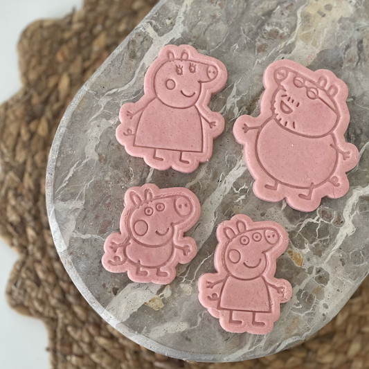 Four pink Peppa Pig shaped items arranged on a gray marbled stone plate, with a brown woven mat in the background.