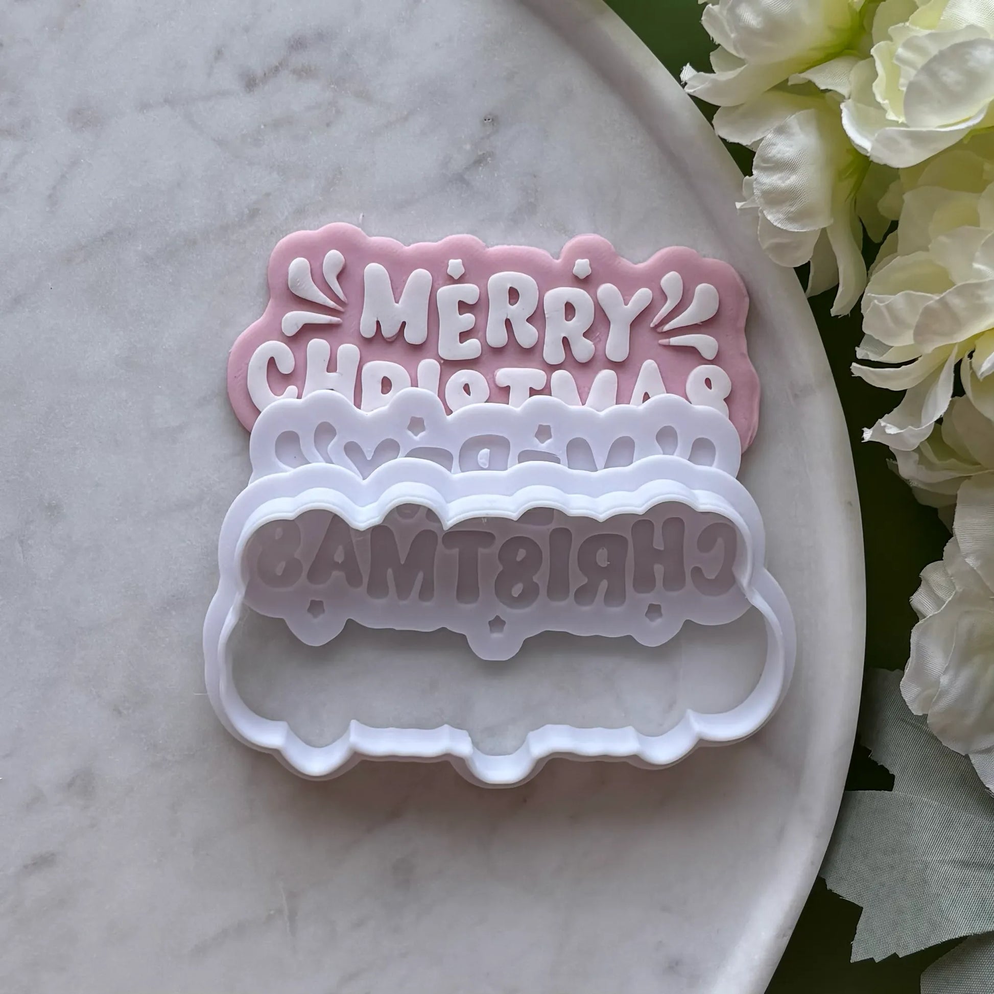 ‘Merry Christmas’ stamped onto pink fondant using a cookie cutter and debosser, displayed on a marble surface