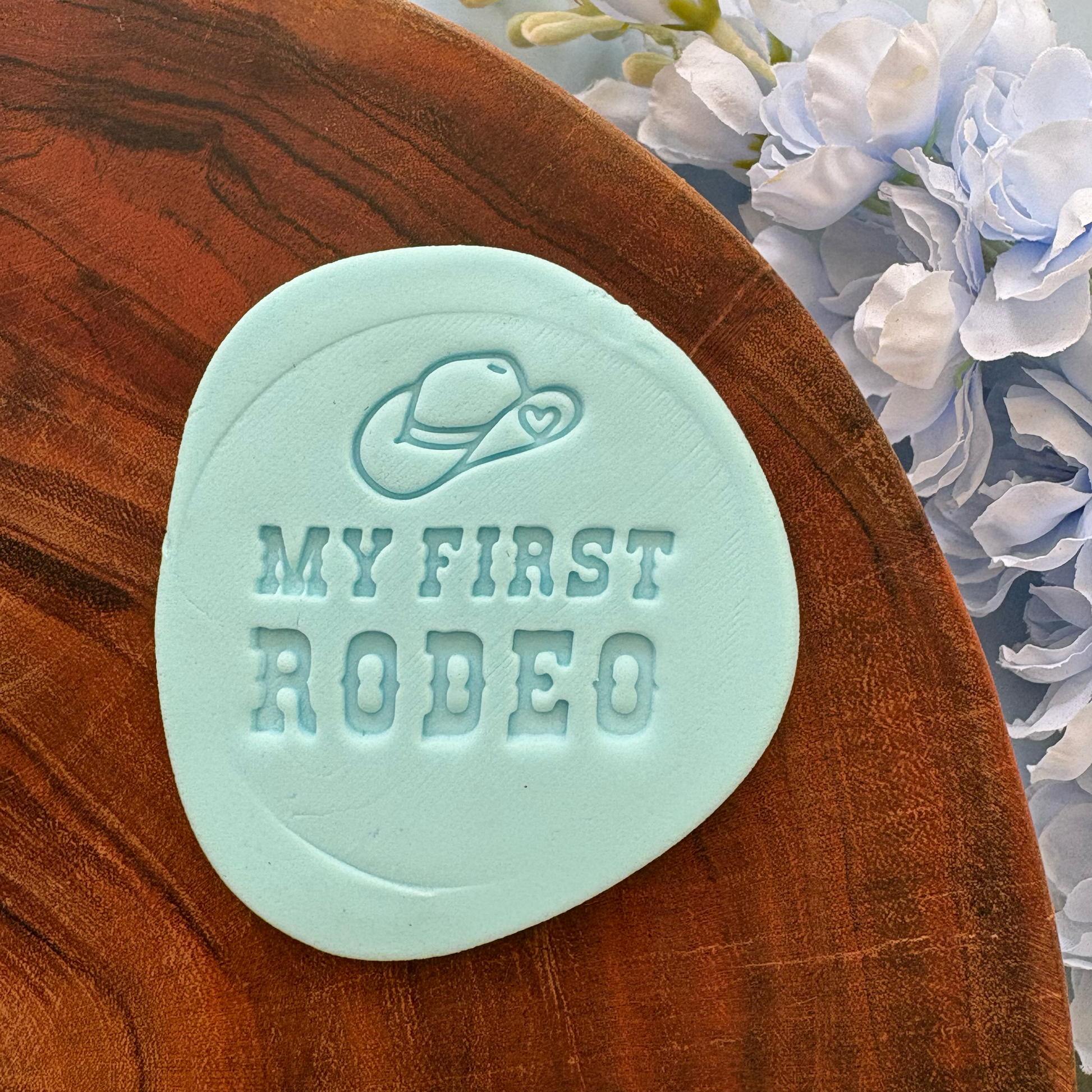 Mint green 'My First Rodeo' stamp on a wooden surface with flowers in the background