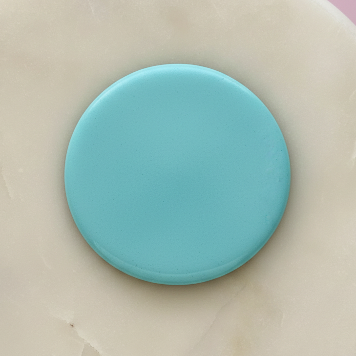 A smooth, circular turquoise blue color swatch resting on a textured beige stone surface with a soft pink background visible in the upper corner.